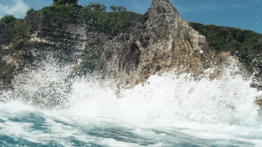 Ocean waves crash against a rocky island at midday causing splashes