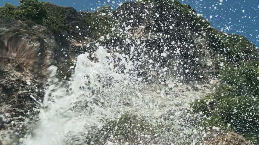 Water splashes dramatically in the foreground with individual droplets visible against a rocky cliff face covered in sparse green vegetation under a clear blue sky