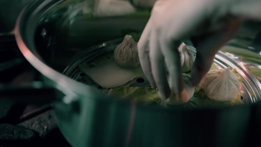 A closeup view of hands carefully placing freshly formed dumplings onto a bed of green leaves within a metal steamer pot ready for cooking