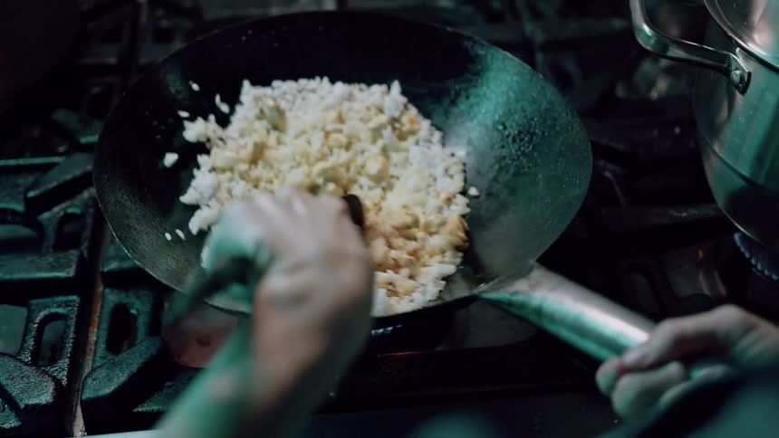 The video captures a closeup of hands actively stirfrying rice in a large wok over a gas burner the rice with visible egg pieces is being tossed and mixed in the hot pan