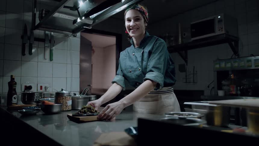 A smiling chef stands in a professional kitchen presenting a freshly prepared dish on a wooden board across the counter