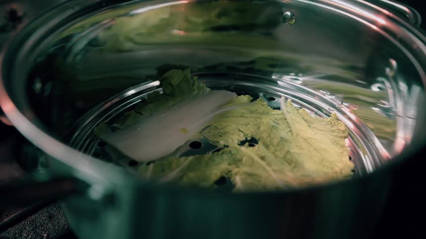 A metal steamer pot contains large light green lettuce leaves cooking in a kitchen setting