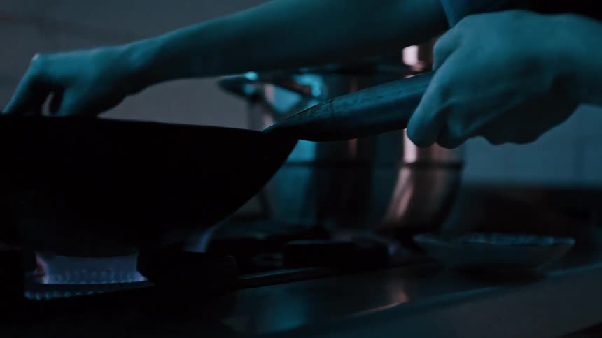 The video shows a closeup of a persons hands holding a black pan over a lit gas burner in the background other kitchen equipment including a pot is visible suggesting a cooking environment