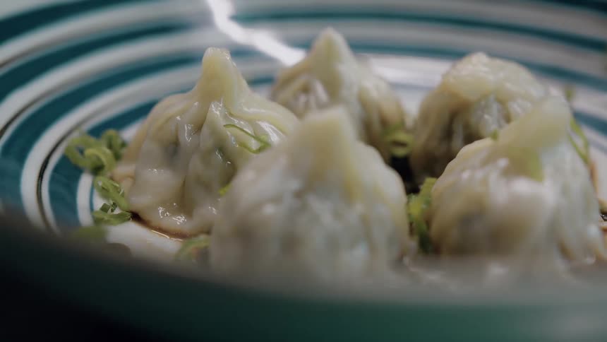 Plump dumplings rest in a striped dish topped with falling sliced scallions and a light drizzle of sauce