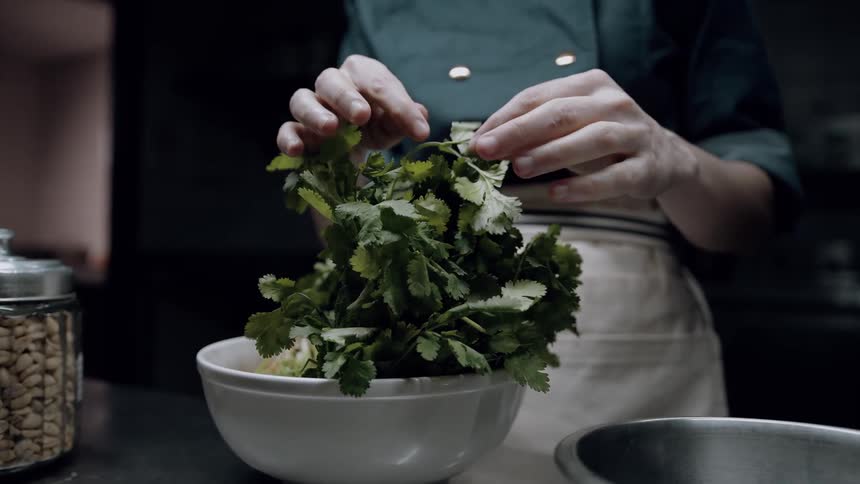 The video shows a chef in a dark green jacket and white apron carefully picking green cilantro leaves from a large bunch in a white bowl in the background theres a metal container filled with what looks like peanuts and other kitchen utensils suggest