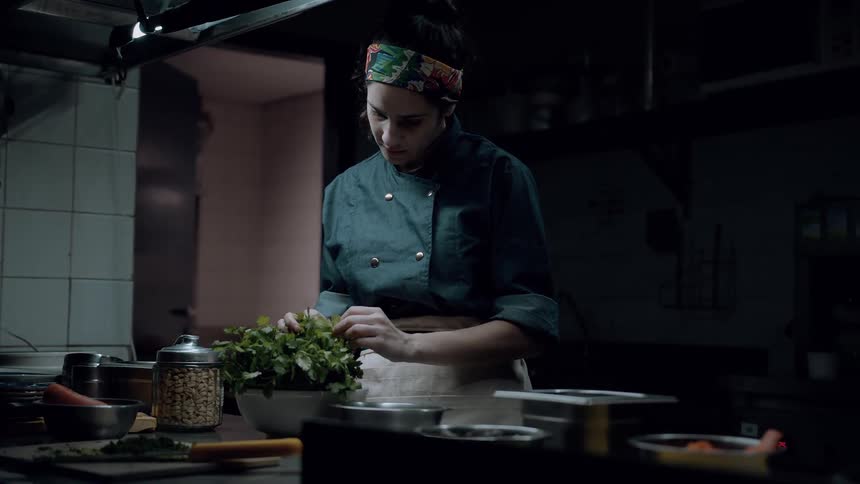 The chef inspects and prepares fresh herbs in a dimly lit kitchen
