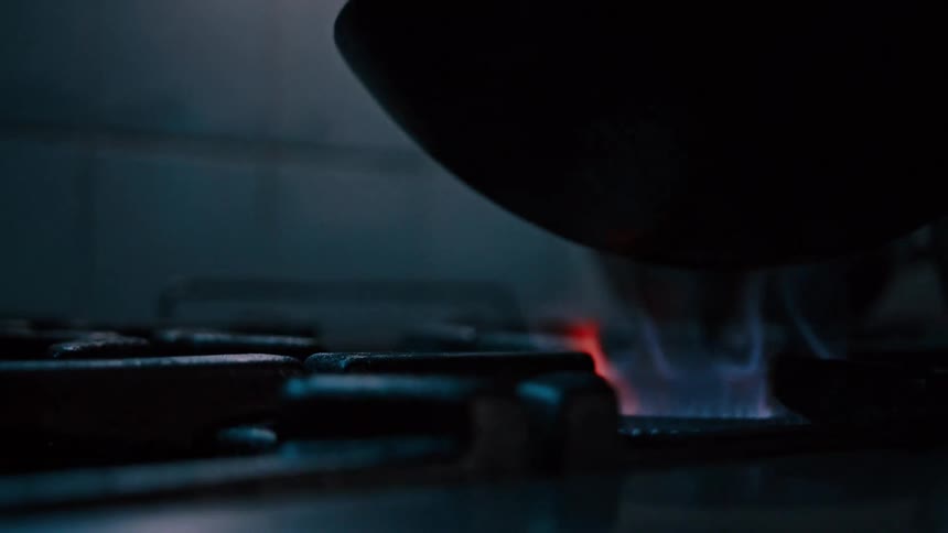 The video shows a dark pan with a subtle blue flame underneath it suggesting a gas burner is lit the pan is empty and rests on a stovetop with a larger reflective metal pot visible in the blurred background