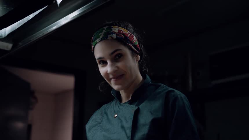 The video shows a woman in a dark chefs jacket and a colorful patterned headband with her hair tied back she looks up and smiles at the camera then looks down again all within a dimly lit kitchen setting