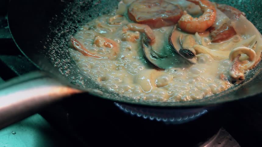 The video shows a closeup of a dark pan containing a creamy sauce with various seafood including shrimp mussels and squid rings simmering over a gas burner the sauce is actively bubbling indicating its hot and in the process of cooking