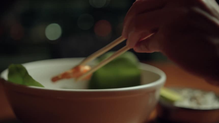 A persons arm reaches to take food from a bowl using chopsticks