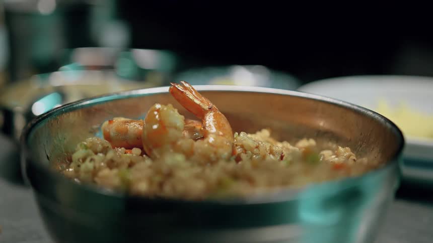 A pair of hands garnishes a metal bowl of fried rice with shrimp adding vegetables and adjusting the presentation with other kitchen items in the blurred background