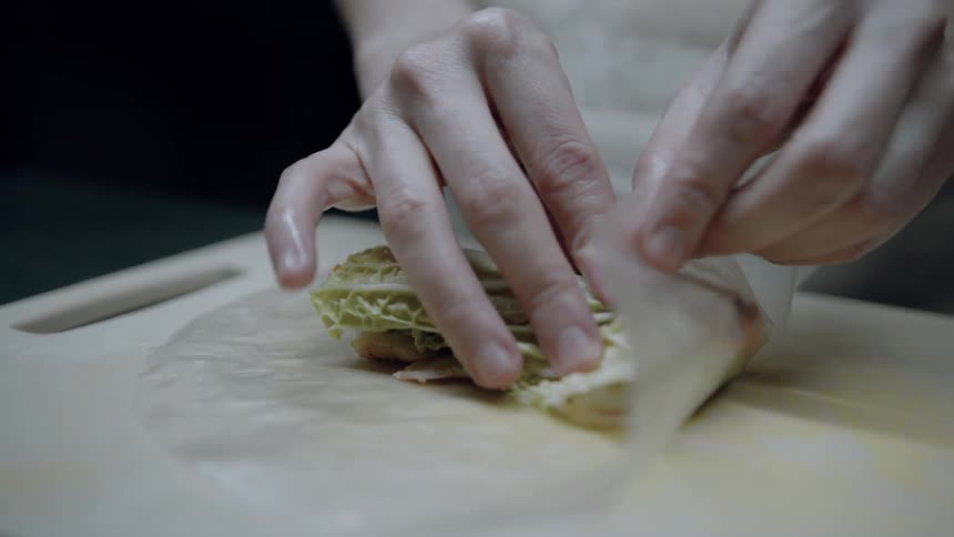 The video shows hands carefully wrapping fresh ingredients including what looks like lettuce and possibly shrimp and carrots into a spring roll wrapper on a wooden cutting board the person then skillfully folds the edges to create a tightly sealed spri