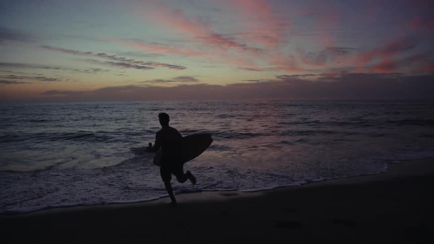 A man holds a surfboard as he runs into the ocean from the beach at sunset