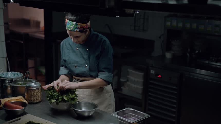 A young female chef prepares vegetables in a bowl while standing in the kitchen