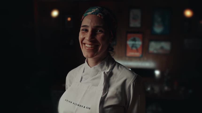 A smiling chef in a white apron stands confidently in a warmly lit restaurant