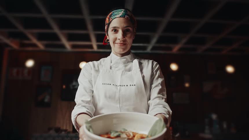 A smiling female chef holds a bowl behind a counter in a warm kitchen