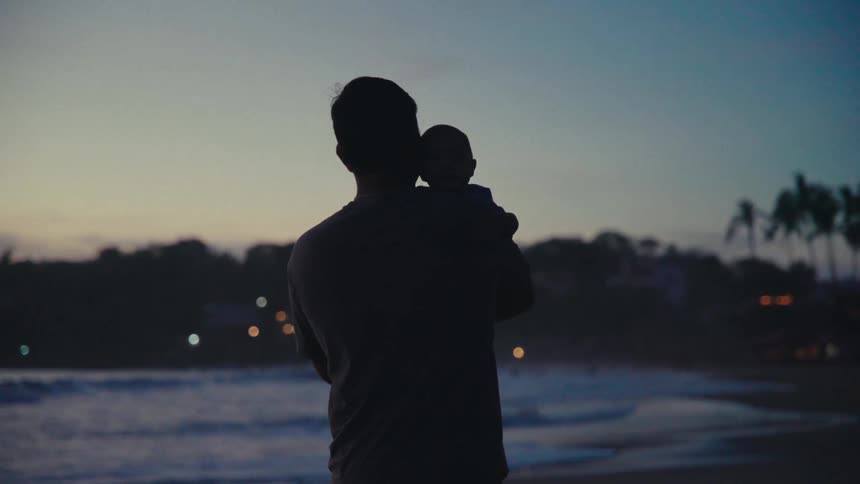 A man holding a baby on his should walking on the beach at sunset