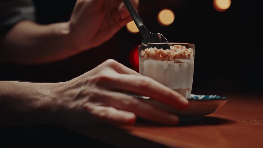 A hand holds a dessert glass while scooping from it under warm ambient lighting