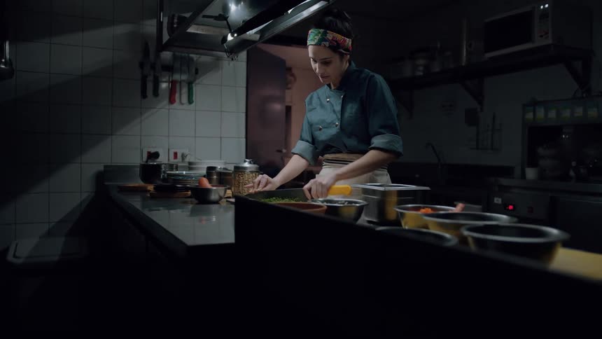 A young chef wearing a dark green chefs coat and a patterned headscarf stands in a dimly lit kitchen chopping green herbs on a cutting board with a yellow handled knife