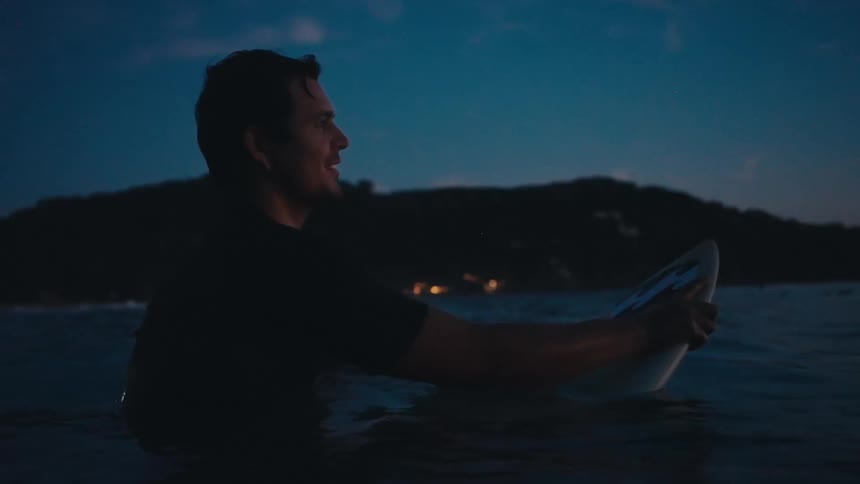 A man on a surfboard in the ocean at dusk looking towards a distant coastline with a serene expression