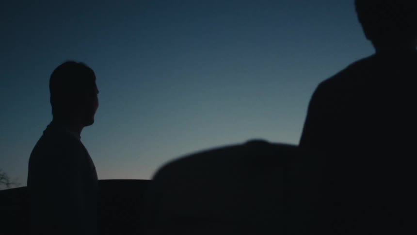 Two people are silhouetted against a fading twilight sky seen from behind as they stand outdoors