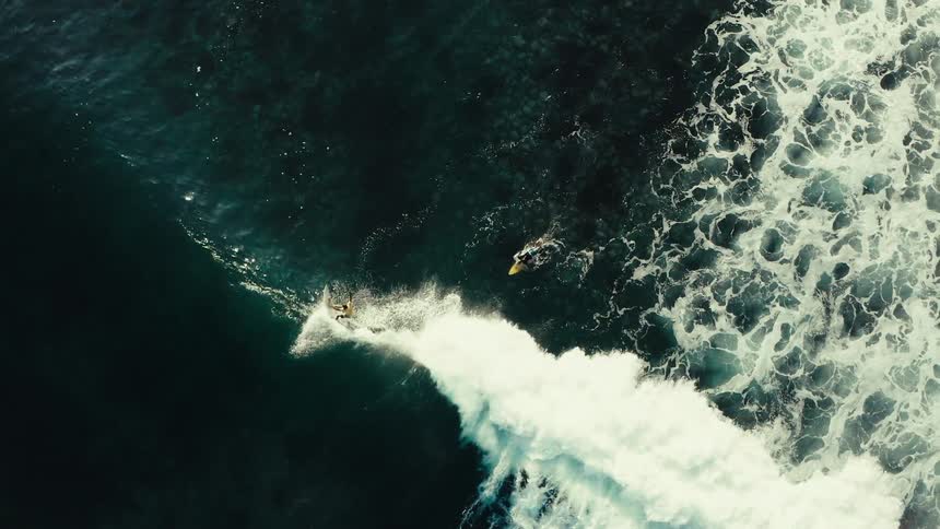 Surfers ride powerful waves as white foam trails across deep ocean waters from above