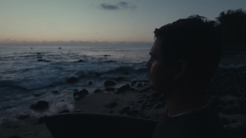 A man sits on a rocky beach at sunset as ocean waves crash against the rocks