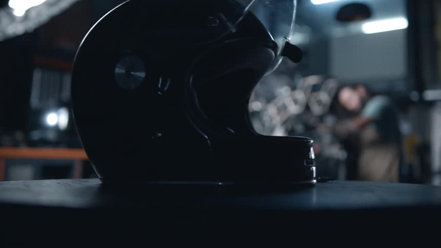 A motorcycle helmet sits on a table in a repair shop while an employee repairs the motorcycle in the background