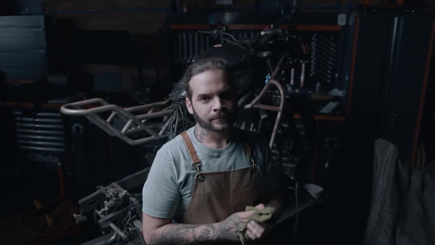 A man wipes his hands while standing in a garage in front of a motorcycle being repaired