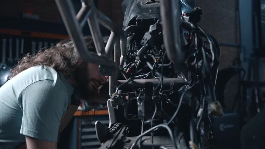 An adult man with long curly hair and a beard wearing a light colored shirt leans in closely to examine the intricate parts of a motorcycle engine in a garage