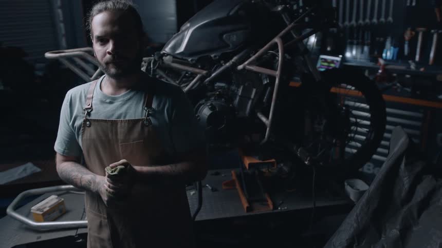 A young male motorcycle mechanic wipes his fingers with a cloth while standing in his workshop