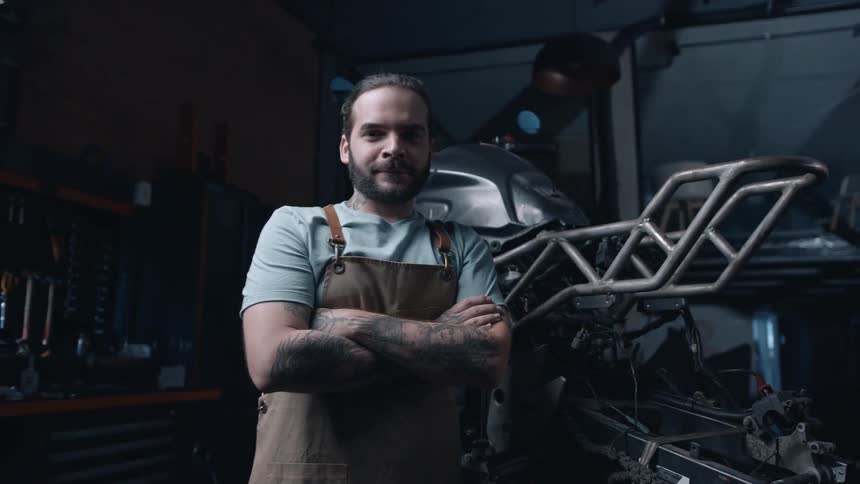 A mechanic smiles while standing next to a motorcycle chassis in a repair shop