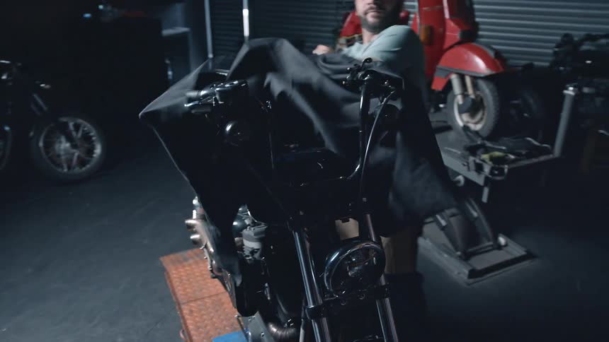A man removes a cover from a motorcycle revealing the bike inside a workshop