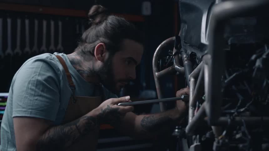 A man with tattoos and a bun is intently working on a motorcycle engine in a dimly lit workshop using a wrench to tighten a part