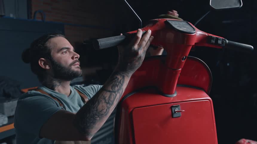 A young mechanic with a beard long hair tied back and tattooed arms wearing an apron works on the handlebars of a bright red scooter in a garage