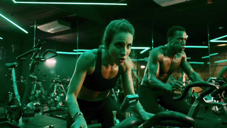 A couple both sweating and showing intense effort ride stationary bikes in a gym with green neon lights