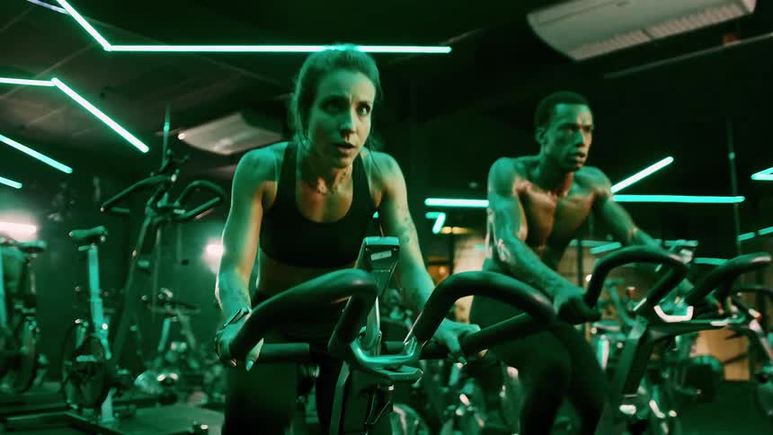 Two athletes push through an intense spin session in a neonlit cycling studio