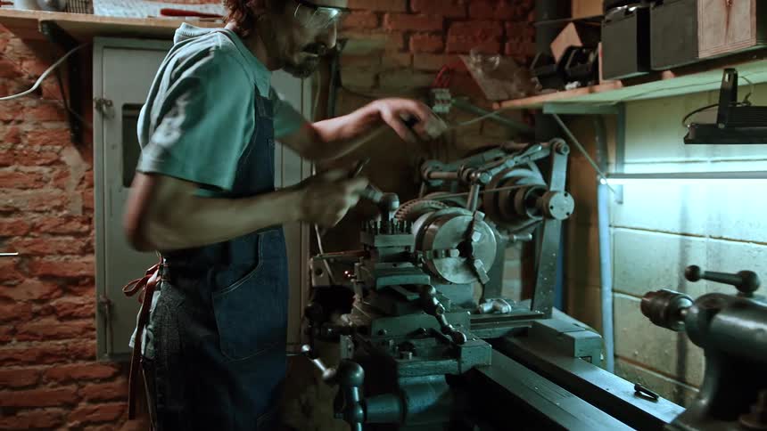 An adult man with long hair glasses and an apron over a light colored shirt works intently on a large metal lathe in a workshop with brick walls and shelves filled with various items