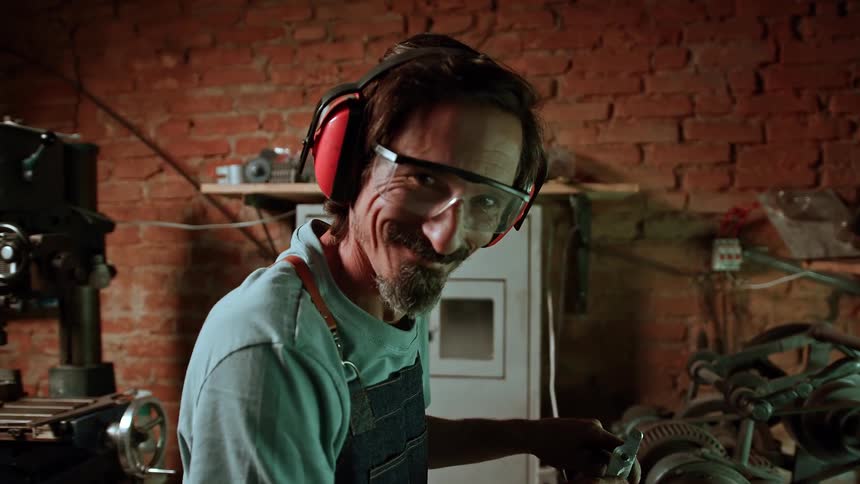 An adult man an artisan with a beard and glasses wearing orange ear protection and an apron over a light colored shirt looks at the viewer and smiles while working with machinery in a workshop with a brick wall