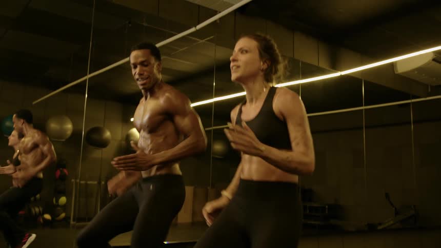 A man with a muscular physique and a woman in athletic wear are performing a dynamic exercise in a gym with large mirrors reflecting their movements and the gyms interior