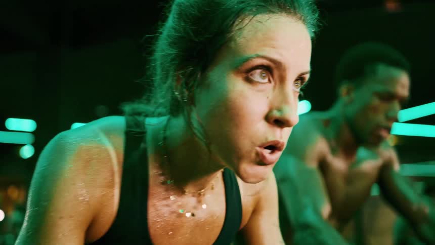 A young adult woman in a black sports bra with sweat visible on her face and shoulders rides a stationary bike with an intense expression in a gym with green neon lighting as another individual rides beside her