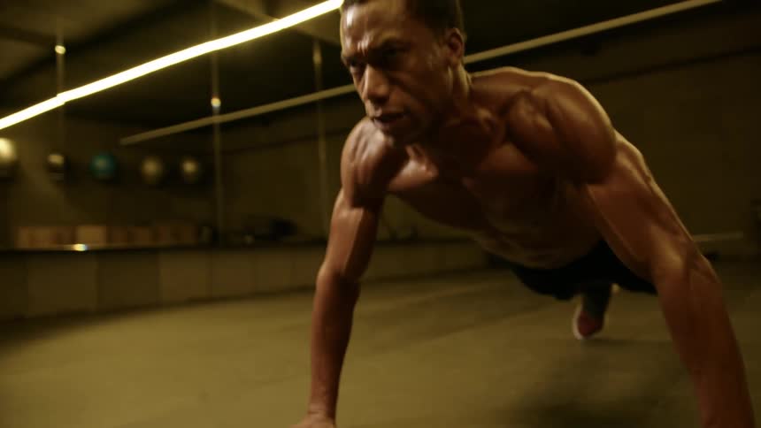 A man with a muscular physique and short hair performs pushups on a dark floor in a gym with horizontal strip lights on the ceiling and a mirrored wall in the background