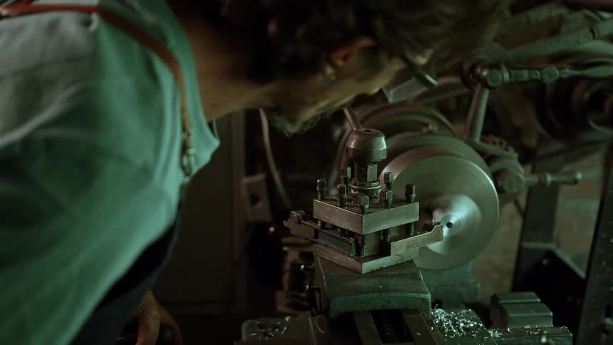 An adult male artisan with a beard and glasses wearing a light colored shirt leans over a large metal lathe machine intently focusing on the metal piece being worked