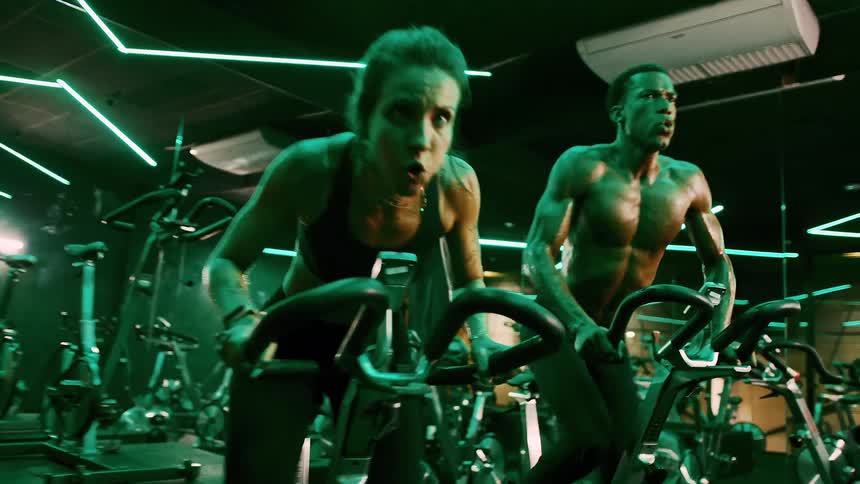 Two adults a woman and a man both covered in sweat and showing intense effort ride stationary bikes side by side in a dimly lit gym with green neon lighting
