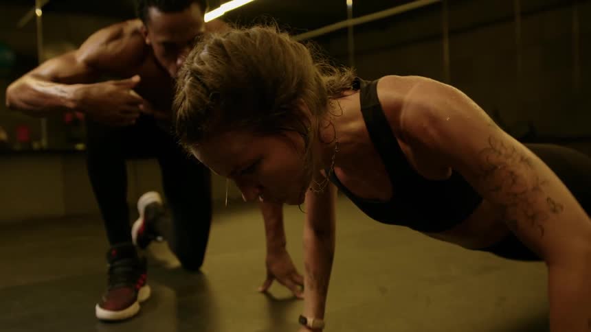 Woman doing pushups with a strained expression as a trainer watches in the background in a dimly lit gym