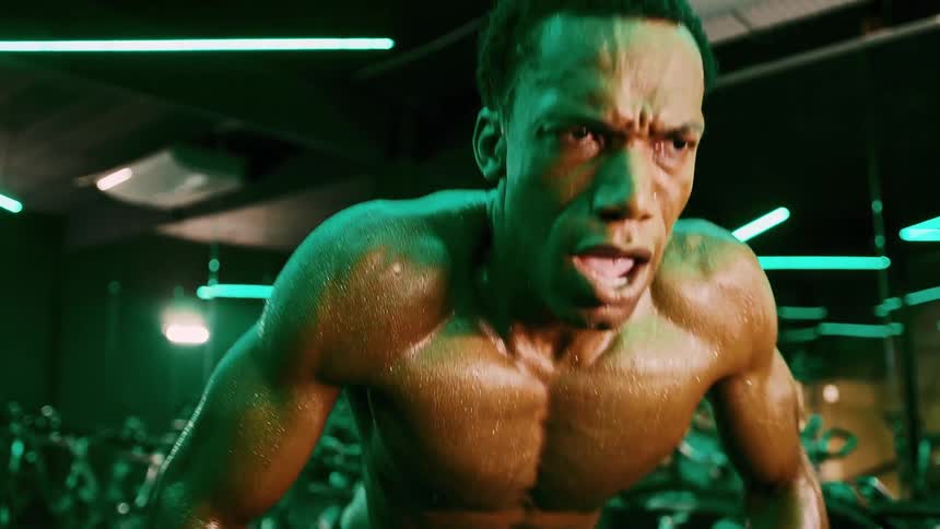 A young adult man with short hair and a muscular build covered in sweat leans forward with a strained expression then breaks into a wide smile in a gym with green lighting