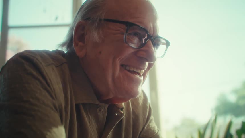 An elderly man celebrates after using a laptop at a table while sitting in living room during daytime