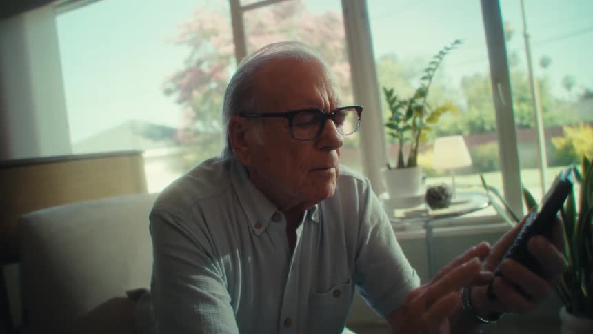 An elderly man sits on a couch while taping on his smartphone screen