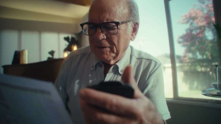 An elderly man sits in his living room looking excited as he reads something on his mobile device