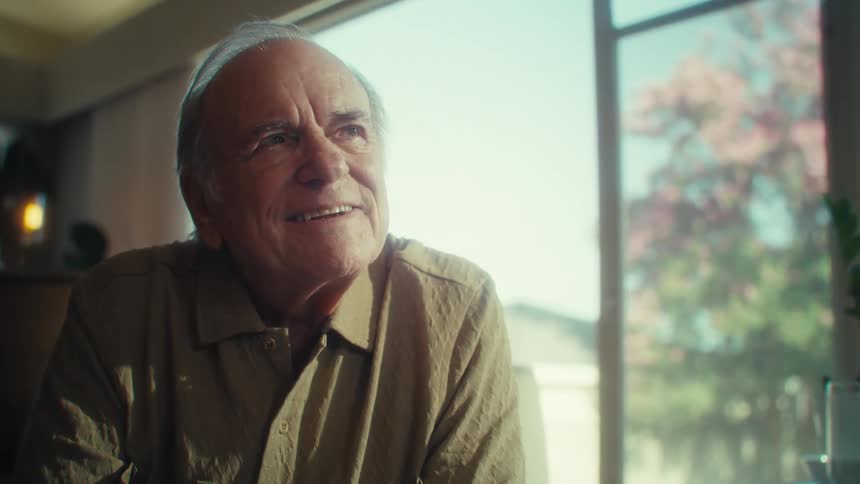 An older adult man with light colored hair wearing a collared shirt and glasses smiles and removes his glasses while looking towards the viewer in a brightly lit room with a large window